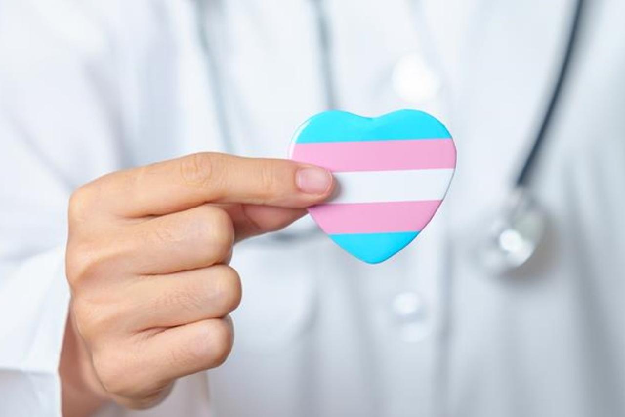 A doctors hand holding a heart shaped pin with the trans flag colours