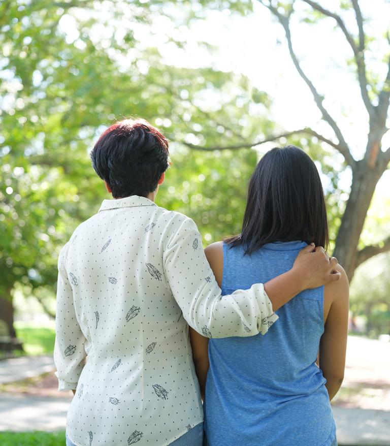 one person with their arm around another person from behind standing in a park with trees in the background